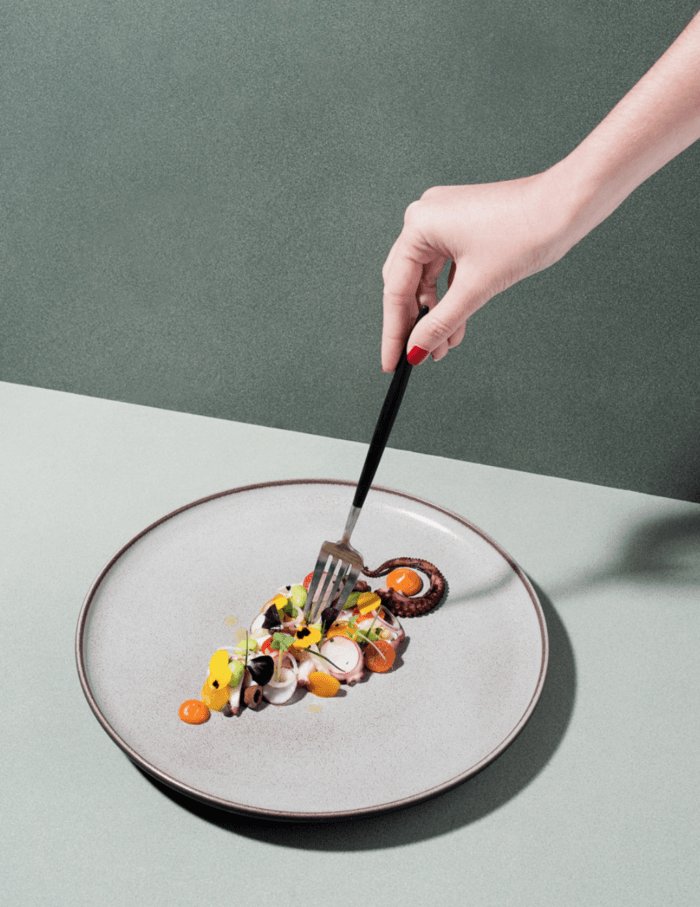 photographie de Caspar Miskin d'une assiette contenant du poulpe et des tomates jaune. une main de femme tiens une fourchette noir